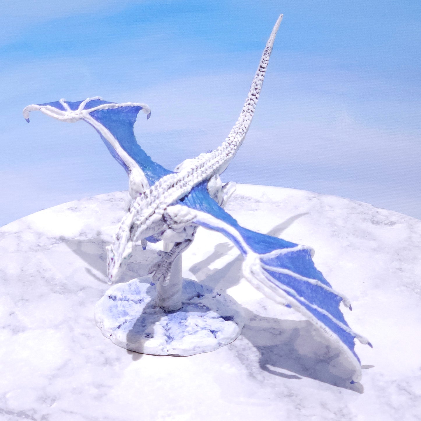A fantasy ice dragon figurine posed on a snowy white surface against a pale blue sky background.