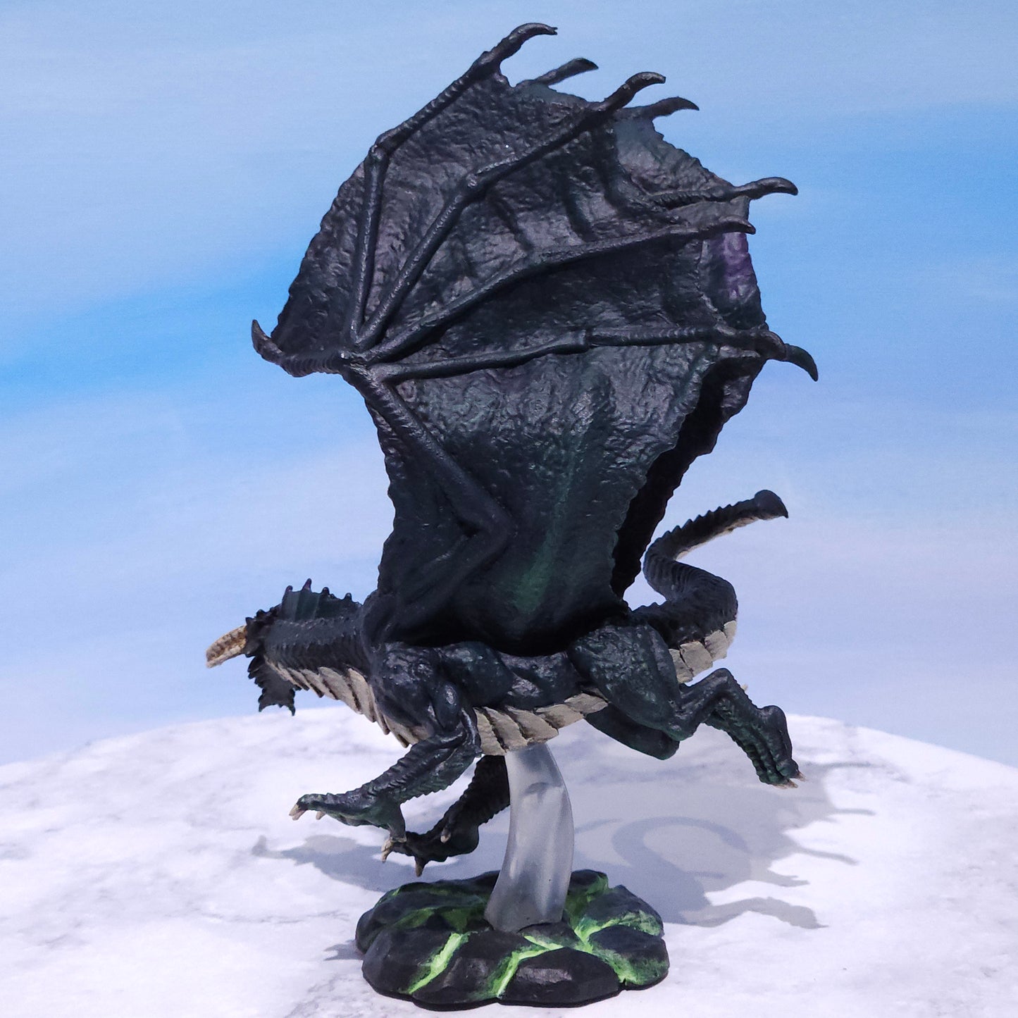 A fantasy dragon figurine perched on a snowy white surface against a blue sky backdrop. The dragon is predominantly black with dark gray wings, small green glowing accents on its body and base, and a clear acrylic display stand.