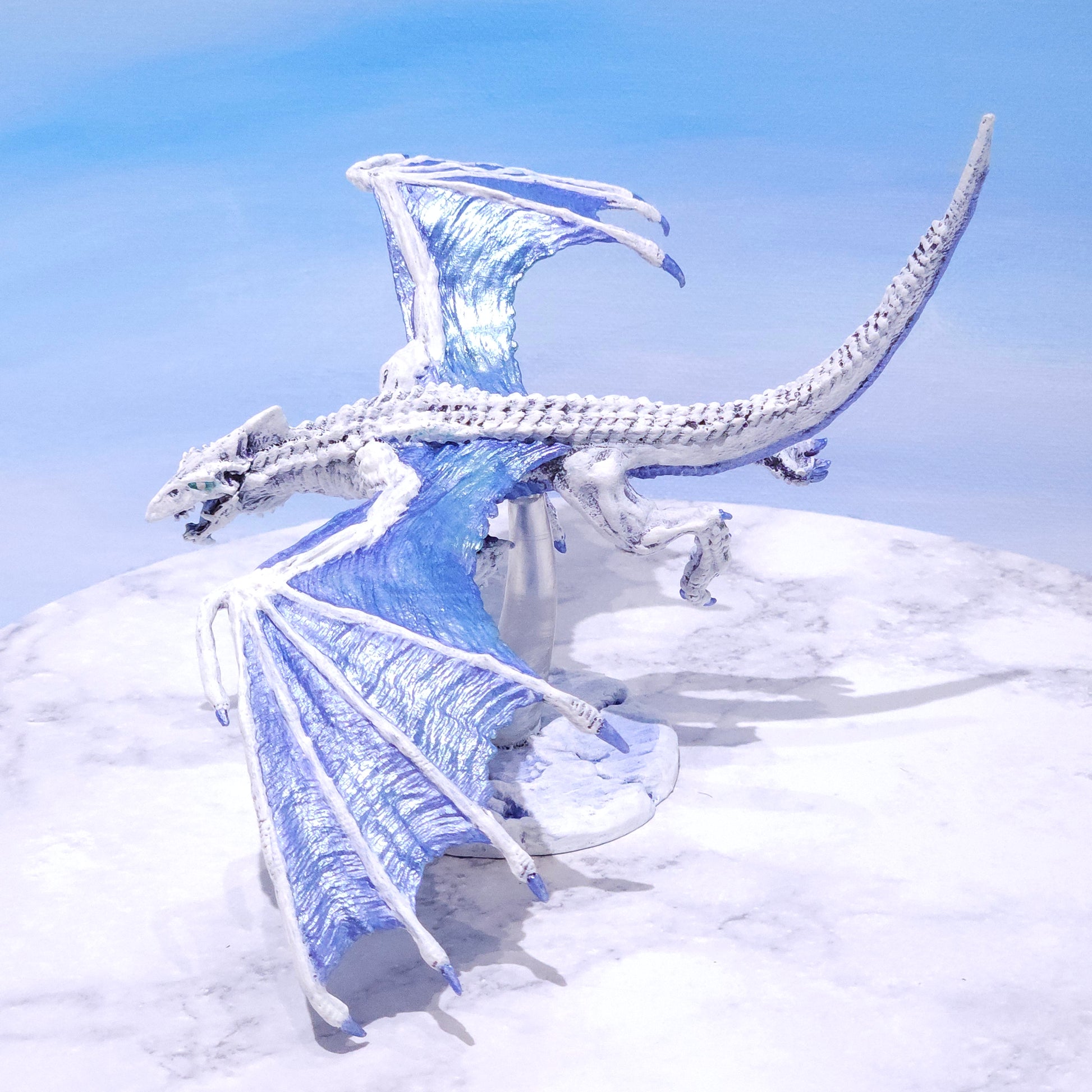 A white and light blue dragon figurine posed against a snowy white wall and pale blue sky background.